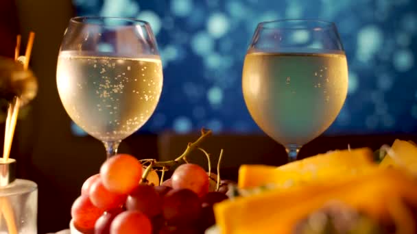 Grapes against bubbles formed inside two wine glass filled with white sparkling wine on the buffet table. Cold beverages showing new year and Christmas celebration theme concept against led television. - Video, Çekim
