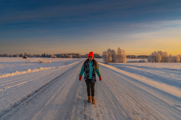 Piękna samotna badaczka w kolorowym ubraniu spacerująca po pustej drodze ze śniegiem pokrytym malowniczym zimowym krajobrazem i światłem słonecznym z horyzontu - Zdjęcie, obraz