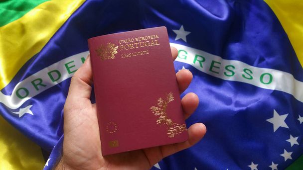 Ανδρικό χέρι κρατώντας πορτογαλικό διαβατήριο με βραζιλιάνικη σημαία στο παρασκήνιο. - Φωτογραφία, εικόνα