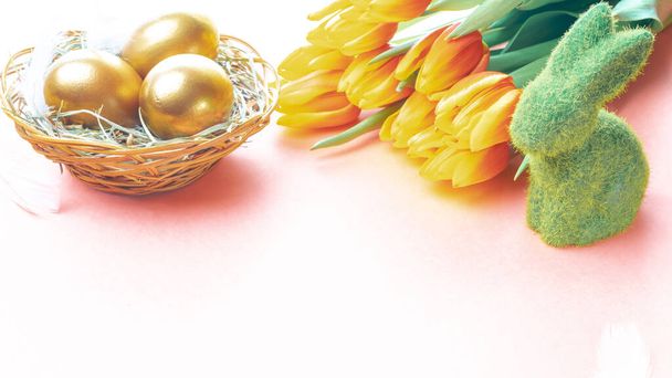 イースターエッグゴールド。春のチューリップのバスケットに黄金の色の卵、ハッピーイースターの装飾でパステルピンクの背景に白い羽。おめでとうございますイースターデザイン - 写真・画像