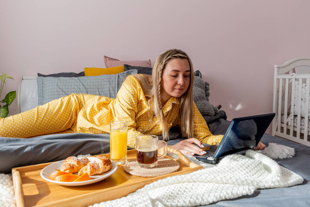 Young woman in yellow pajamas has healthy breakfast and work laptop while lying on bed in bedroom. Home wellbeing concept. Emotional health of a young woman. Wellness - Photo, Image