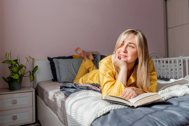 Νεαρή γυναίκα με κίτρινες πιτζάμες διαβάζει βιβλίο ενώ ξαπλώνει στο κρεβάτι στην κρεβατοκάμαρα. Η έννοια της ευημερίας. Συναισθηματική υγεία μιας νεαρής γυναίκας - Φωτογραφία, εικόνα