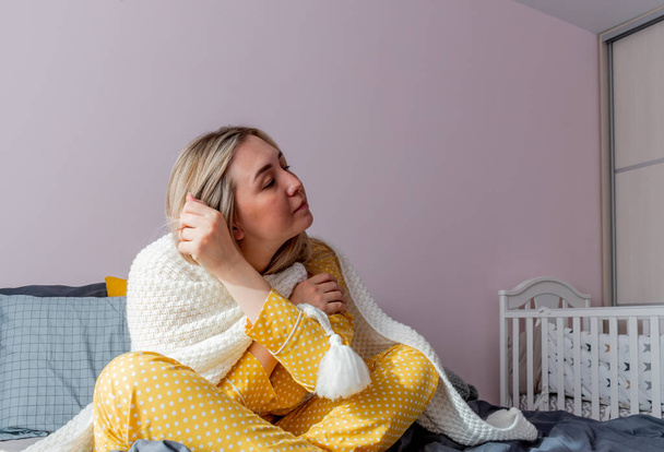 Young woman is wrapped in white plaid while sitting on bed in bedroom. Wellness and mental health concept. Calm pastel colors. - Photo, Image