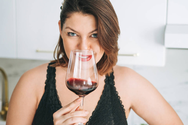 Mooie gelukkig sensuele brunette jonge vrouw plus size lichaam positief met glas rode wijn genieten van haar leven in het huis - Foto, afbeelding