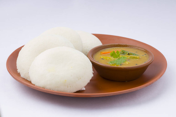 Idly of Idli, Zuid-Indiase belangrijkste ontbijt item dat prachtig is gerangschikt in een aardewerk met een kleine kom van sambar geplaatst op witte achtergrond. - Foto, afbeelding