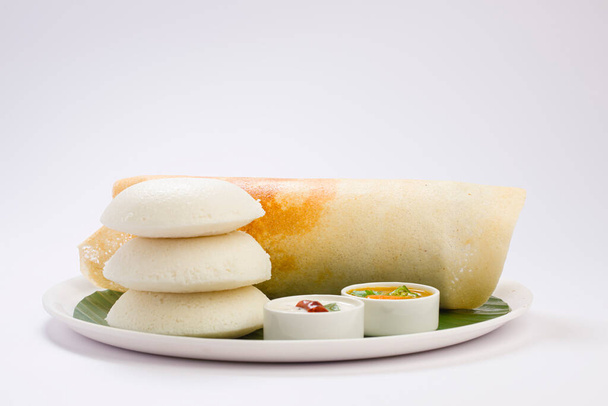 Ghee roast Dosa e Idlii, plato principal de desayuno del sur de la India que está bellamente dispuesto en un plato blanco forrado con hoja de plátano y curry como sambar y chutney sobre fondo blanco. - Foto, Imagen
