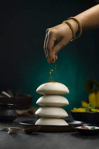 Idly of Idli, Zuid-Indiase belangrijkste ontbijt item dat is geplaatst in een hoop een over andere prachtig en een vrouw hand garnering idli met chili vlokken die in een zwarte plaat op donkere textuur met keuken achtergrond. - Foto, afbeelding