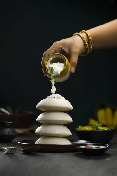Idly or Idli, south-indian main breakfast item which is placed in a heap one over other beautifully and a woman hand pouring some coconut chutney on idly which is in a black plate on dark green texture, kitchen background. - Фото, изображение