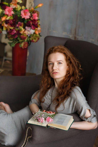 Ritratto di una giovane attraente donna dai capelli rossi che legge un libro su un'accogliente sedia grigia. C'è un vaso di fiori nelle vicinanze. - Foto, immagini