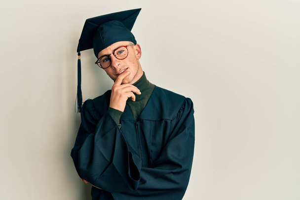 Young caucasian man wearing graduation cap and ceremony robe looking confident at the camera with smile with crossed arms and hand raised on chin. thinking positive.  - Photo, image