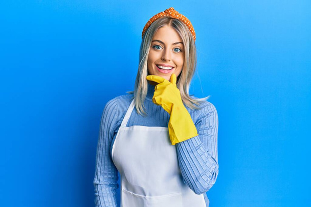 Beautiful blonde woman wearing cleaner apron and gloves looking confident at the camera smiling with crossed arms and hand raised on chin. thinking positive.  - Photo, Image