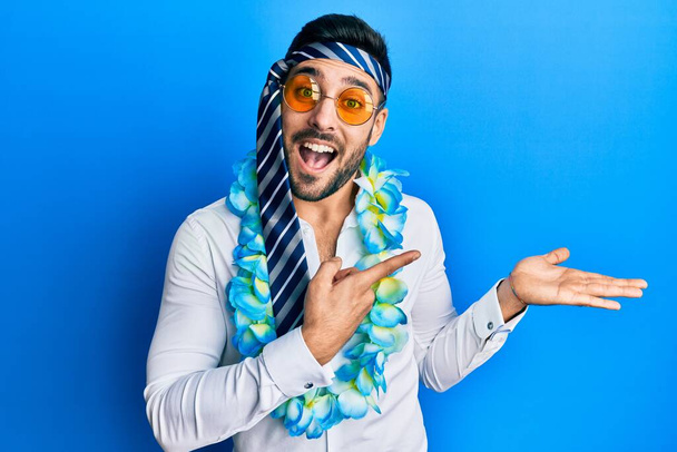 Jeune homme d'affaires hispanique portant fête style drôle avec cravate sur la tête étonné et souriant à la caméra tout en présentant avec la main et pointant du doigt.  - Photo, image
