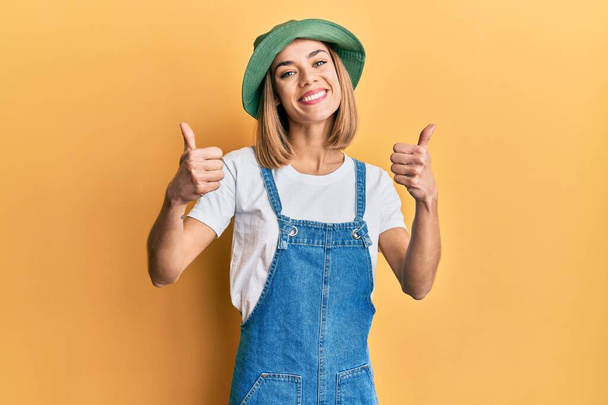 Young caucasian blonde woman wearing denim jumpsuit and hat with 90s style success sign doing positive gesture with hand, thumbs up smiling and happy. cheerful expression and winner gesture.  - Photo, Image