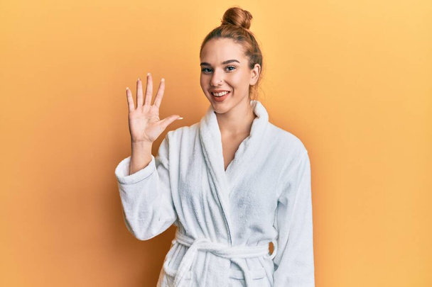 Junge blonde Frau im Bademantel zeigt mit Finger Nummer fünf nach oben und lächelt dabei selbstbewusst und glücklich.  - Foto, Bild