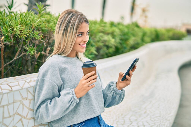 Jovencita rubia usando smartphone y tomando café en el parque. - Foto, imagen