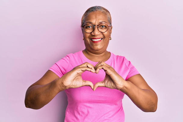 Donna afroamericana anziana che indossa abiti casual e occhiali sorridenti innamorati che mostrano il simbolo del cuore e la forma con le mani. concetto romantico.  - Foto, immagini