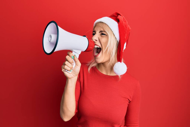 Jeune femme blonde portant un chapeau de Noël criant à l'aide d'un mégaphone sur fond rouge isolé. - Photo, image