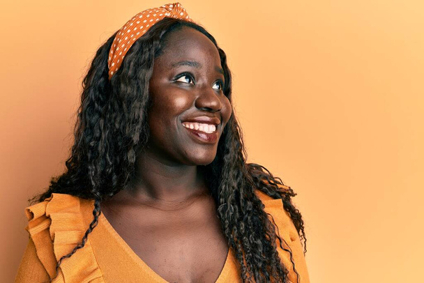 Afrykańska młoda kobieta ubrana w luźne ubrania na żółtym tle patrząc z uśmiechem na twarzy, naturalnym wyrazem twarzy. śmiać się pewnie.  - Zdjęcie, obraz