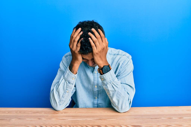 Junge gut aussehende afrikanisch-amerikanische Mann in lässiger Kleidung auf dem Tisch sitzend und unter Kopfschmerzen verzweifelt und gestresst, weil Schmerzen und Migräne. Hände auf den Kopf.  - Foto, Bild