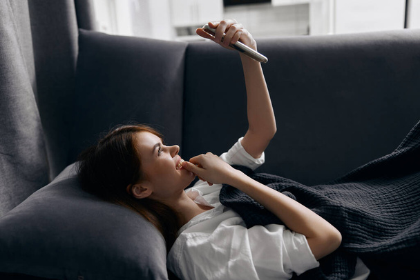 όμορφη γυναίκα με ένα τηλέφωνο στα χέρια της βρίσκεται σε έναν καναπέ μέσα στο σπίτι - Φωτογραφία, εικόνα