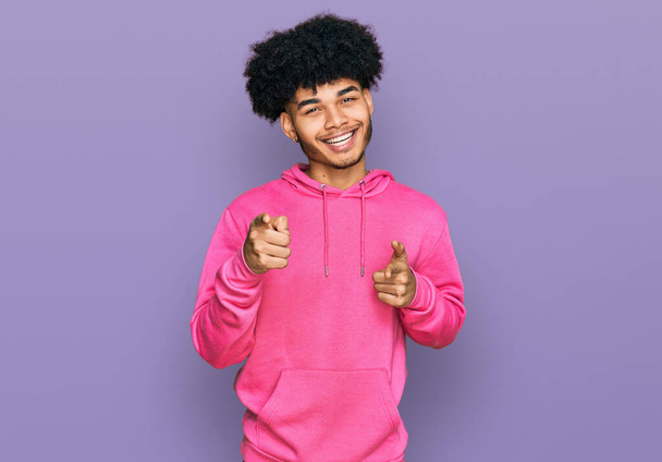 カジュアルなピンクのスウェットシャツを着たアフロヘアの若いアフリカ系アメリカ人男性が、楽しそうで面白い顔をしたカメラを指差している。良いエネルギーと振動は.  - 写真・画像
