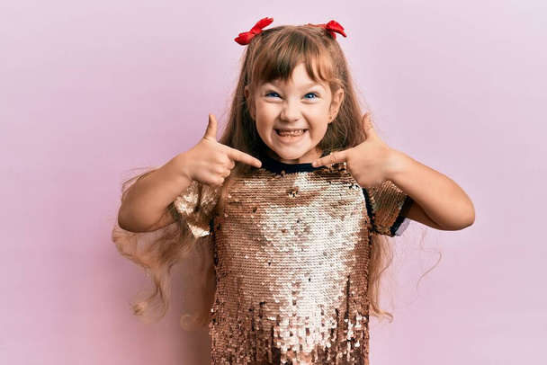 Kleines kaukasisches Mädchen in festlichem Paillettenkleid sieht selbstbewusst aus, mit einem Lächeln im Gesicht, und zeigt stolz und glücklich mit den Fingern.  - Foto, Bild