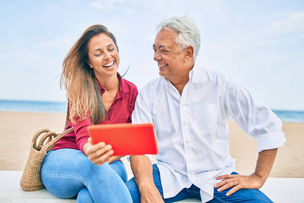 Middle age hispanic couple using touchpad sitting on the bench at the beach. - Zdjęcie, obraz