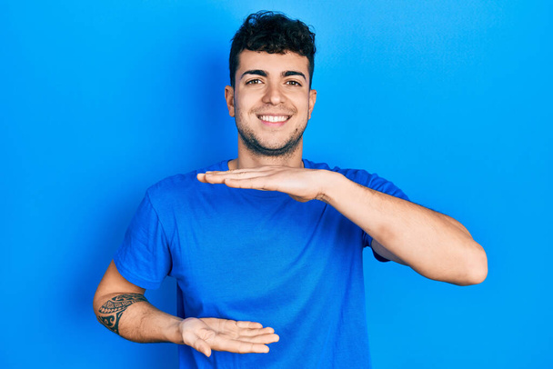Mladý hispánský muž v neformální modré tričko gestikuluje s rukama ukazujícími velkou a velkou značku velikosti, symbol míry. s úsměvem na kameru. koncepce měření.  - Fotografie, Obrázek