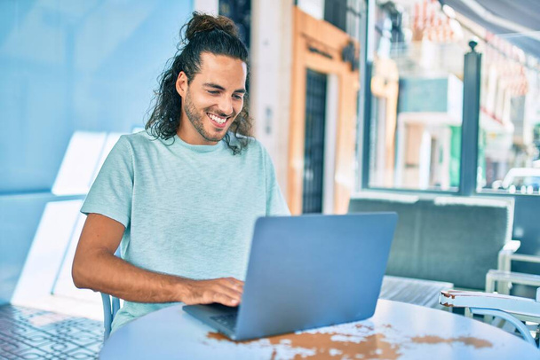 Genç İspanyol gülümseyen adam kahve dükkanının terasında dizüstü bilgisayarla çalışırken mutlu oluyor.. - Fotoğraf, Görsel