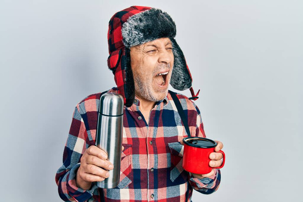 Bello tuttofare maturo che indossa un cappello invernale con paraorecchie che beve caffè caldo da termo arrabbiato e pazzo urlando frustrato e furioso, urlando di rabbia. rabbia e concetto aggressivo.  - Foto, immagini