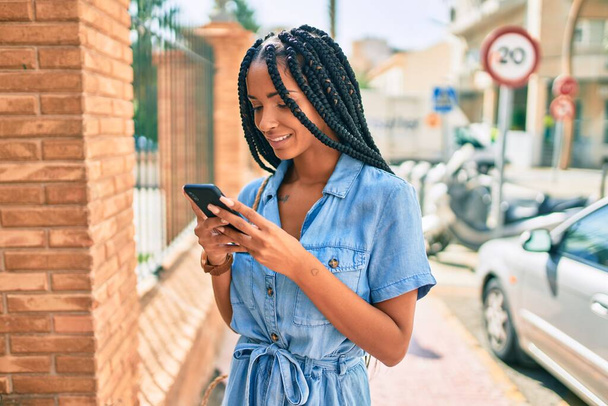 Giovane donna afroamericana sorridente felice utilizzando smartphone in città. - Foto, immagini