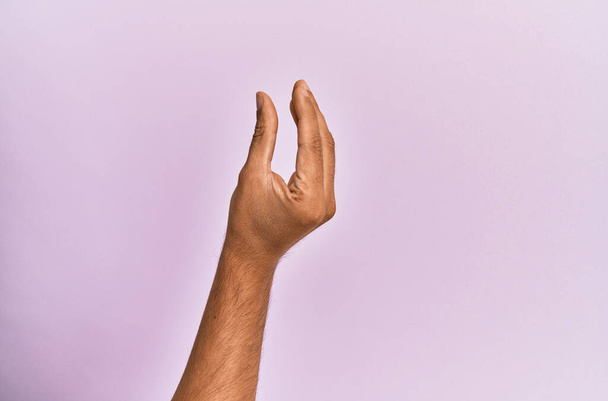 Brazo y mano del joven caucásico sobre fondo aislado rosa recogiendo y tomando cosa invisible, sosteniendo el objeto con los dedos mostrando espacio  - Foto, Imagen