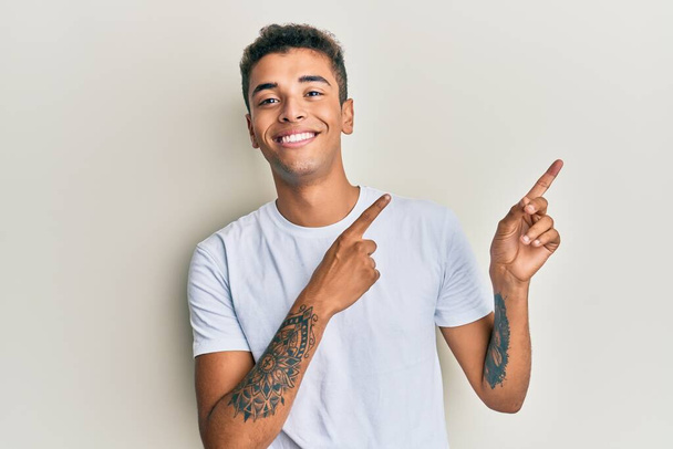Young handsome african american man wearing casual white tshirt smiling and looking at the camera pointing with two hands and fingers to the side.  - Fotografie, Obrázek