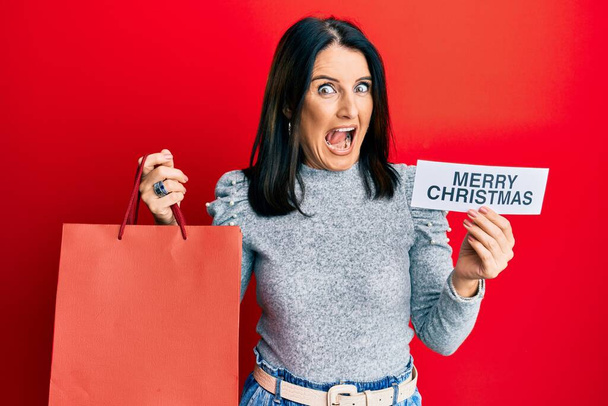 Брюнетка среднего возраста с веселым рождественским посланием и сумкой для покупок празднует безумие и удивляется успеху с открытыми глазами, крича взволнованно.  - Фото, изображение