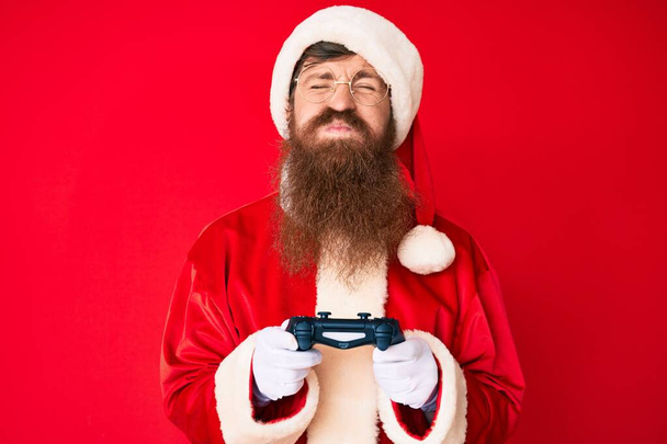 Schöner junger roter Mann mit langem Bart im Weihnachtsmann-Kostüm, der Videospiele spielt und Wangen mit lustigem Gesicht aufbläst. Mund mit Luft aufgeblasen, Luft einfangen.  - Foto, Bild