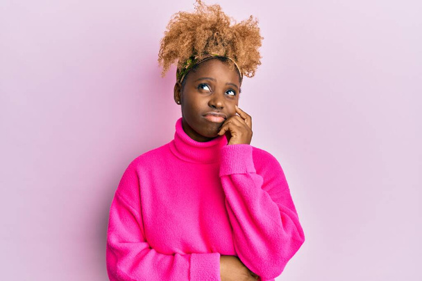 Jeune femme africaine aux cheveux afro portant un pull d'hiver décontracté pensant concentré sur le doute avec le doigt sur le menton et regardant vers le haut se demandant  - Photo, image