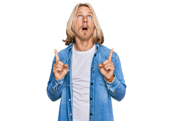 Caucasian man with blond long hair wearing casual denim jacket amazed and surprised looking up and pointing with fingers and raised arms.  - Photo, Image