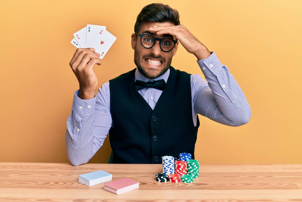 bel homme croupier hispanique assis sur la table avec des jetons de poker et des cartes stressé et frustré avec la main sur la tête, visage surpris et en colère  - Photo, image