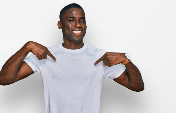 Mladý africký Američan nosí ležérní bílé tričko vypadá sebevědomě s úsměvem na tváři, ukazuje se s prsty hrdý a šťastný.  - Fotografie, Obrázek