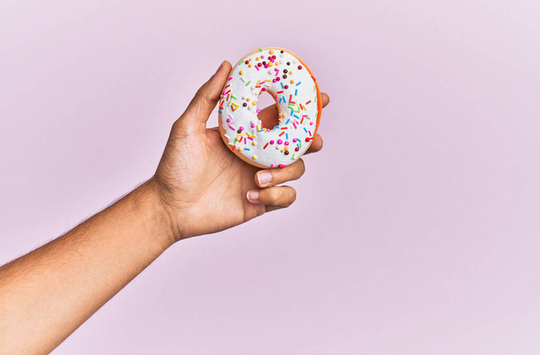Hand of hispanic man holding donut over isolated pink background. - Foto, afbeelding