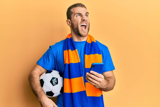 Молодой кавказский мужчина, держащий футбольный мяч, глядя на смартфон злой и безумный кричащий расстроенный и разъяренный, кричащий от злости, глядя вверх.  - Фото, изображение