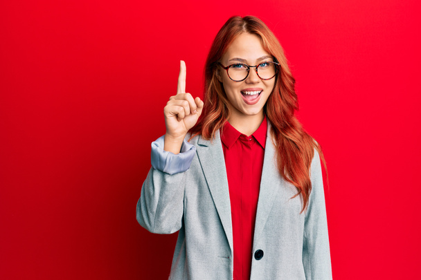 Young redhead woman wearing business jacket and glasses pointing finger up with successful idea. exited and happy. number one.  - Photo, Image