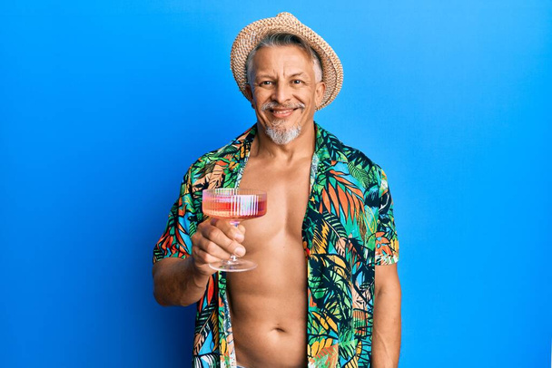 Чоловік середнього віку сіро-волохатий в літньому стилі п'є коктейль, виглядає позитивно і щасливо стоячи і посміхаючись з впевненою посмішкою, показуючи зуби
  - Фото, зображення