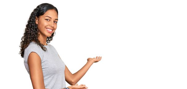 Młoda afrykańska dziewczyna ubrana w luźne ubrania zapraszając do wejścia uśmiechnięty naturalny z otwartą ręką  - Zdjęcie, obraz