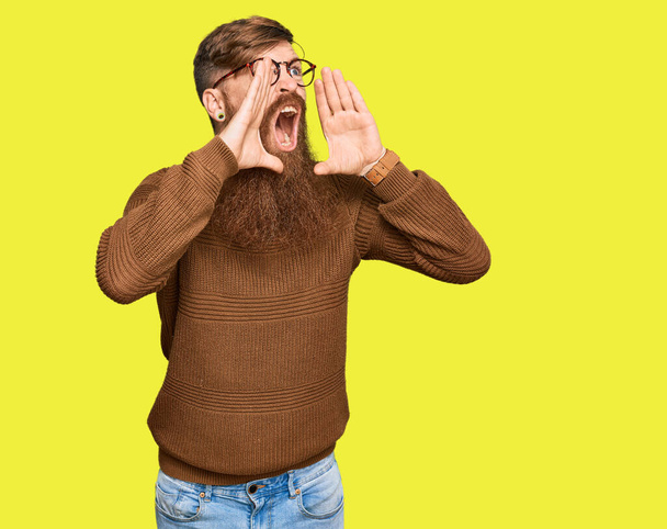 Молодой ирландский рыжий мужчина в повседневной одежде и очках кричит сердито, с руками над ртом  - Фото, изображение