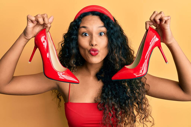 Mujer latina joven sosteniendo zapatos rojos de tacón alto mirando a la cámara soplando un beso siendo encantador y sexy. expresión de amor.  - Foto, imagen