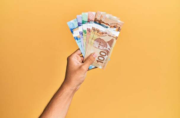 Hand of hispanic man holding canadian dollars over isolated yellow background. - Foto, Imagen