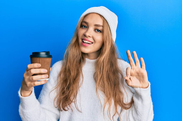 Schöne blonde kaukasische Frau, die eine Tasse Kaffee zum Mitnehmen trinkt, tut ok Zeichen mit den Fingern, lächelt freundlich gestikulierend hervorragendes Symbol  - Foto, Bild