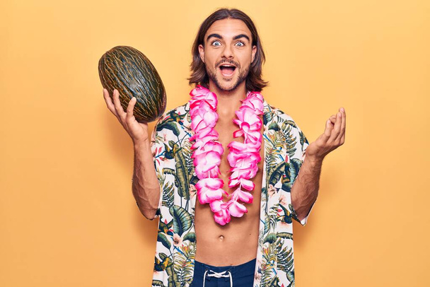 Молодой красивый мужчина в купальниках и гавайских леях, держа дыню, празднуя достижение со счастливой улыбкой и выражением победителя с поднятой рукой  - Фото, изображение