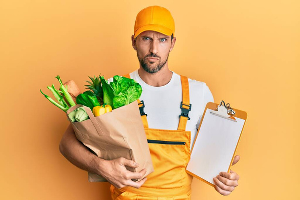 Young handsome man wearing courier uniform with groceries and clipboard skeptic and nervous, frowning upset because of problem. negative person.  - Fotoğraf, Görsel
